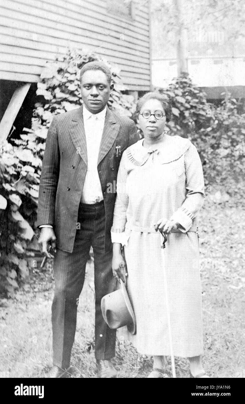 African American husband and wife standing next to each other in front of a wood building and shrubbery, the woman is wearing a light-colored shirt and long skirt and is holding a hat in one hand and a cane in the other, the man is wearing a full dark-colored suit with a light colored shirt underneath and he is holding a cigar in one hand, 1950. Stock Photo