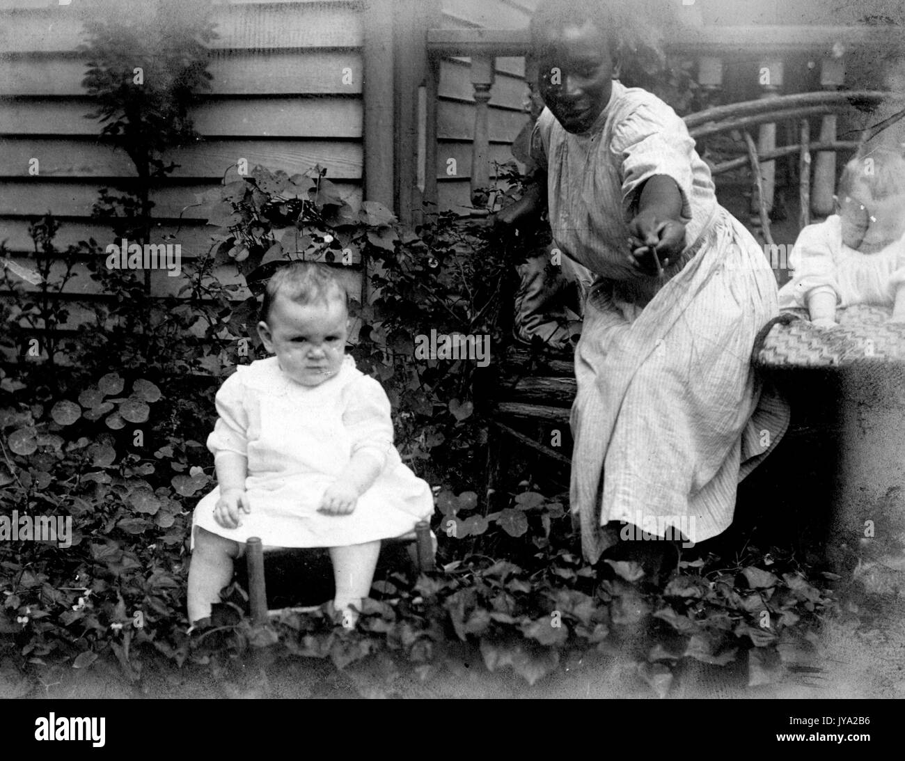 African-American nanny in a garden beside a home, wearing a plain dress and holding out a flower, with two young Caucasian babies, 1910. Stock Photo