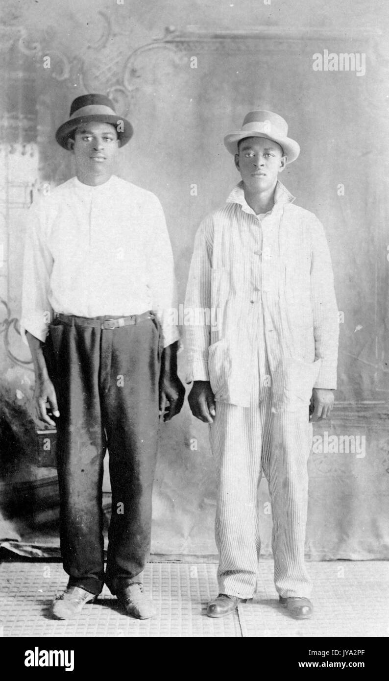 Two African-American brothers standing side by side and posing for a studio portrait, the younger brother wearing a white suit, the older brother wearing a white shirt and dark pants, both wearing hats, with hands at their sides, blank facial expressions, 1910. Stock Photo
