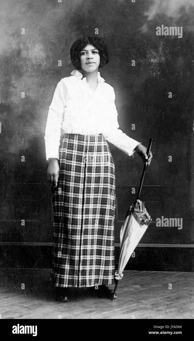 Young African American woman wearing a white blouse and checkered skirt, holding an umbrella and posing for a photograph in a portrait studio, 1932. Stock Photo