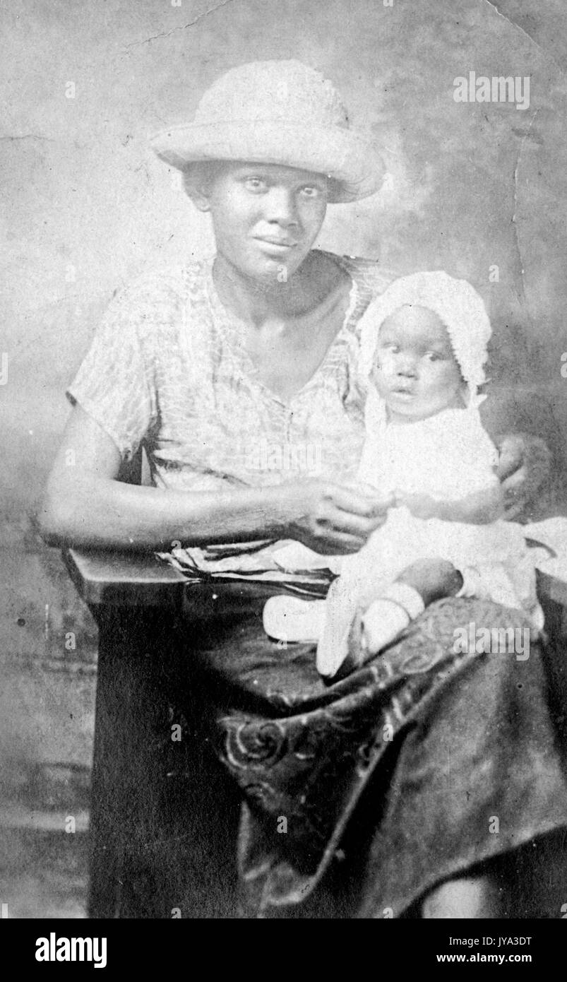 African American mother posing with her child, wearing a wide brimmed hat and with a happy facial expression, the child sitting on her lap and wearing a white dress and bonnet, 1932. Stock Photo