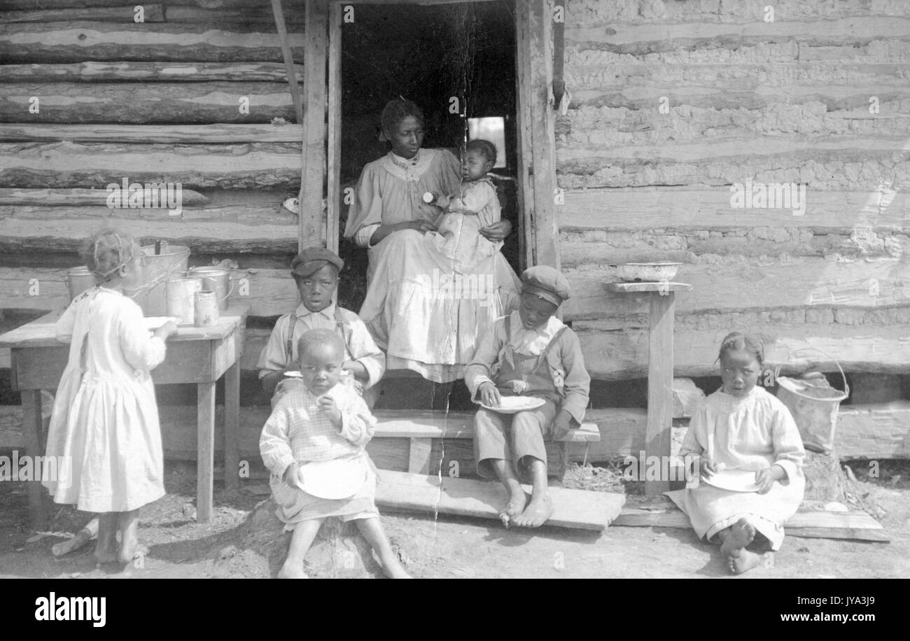 Family of african-american sharecroppers, the mother sitting in the door of a log cabin and holding young baby, several other children sitting and standing on the broken steps of the cabin and eating a meal, using their hands to eat food from bowls, each child wearing a simple dress or overalls, one girl standing at a table to the left and helping herself to food, dusty ground underneath, all children barefoot, 1910. Stock Photo