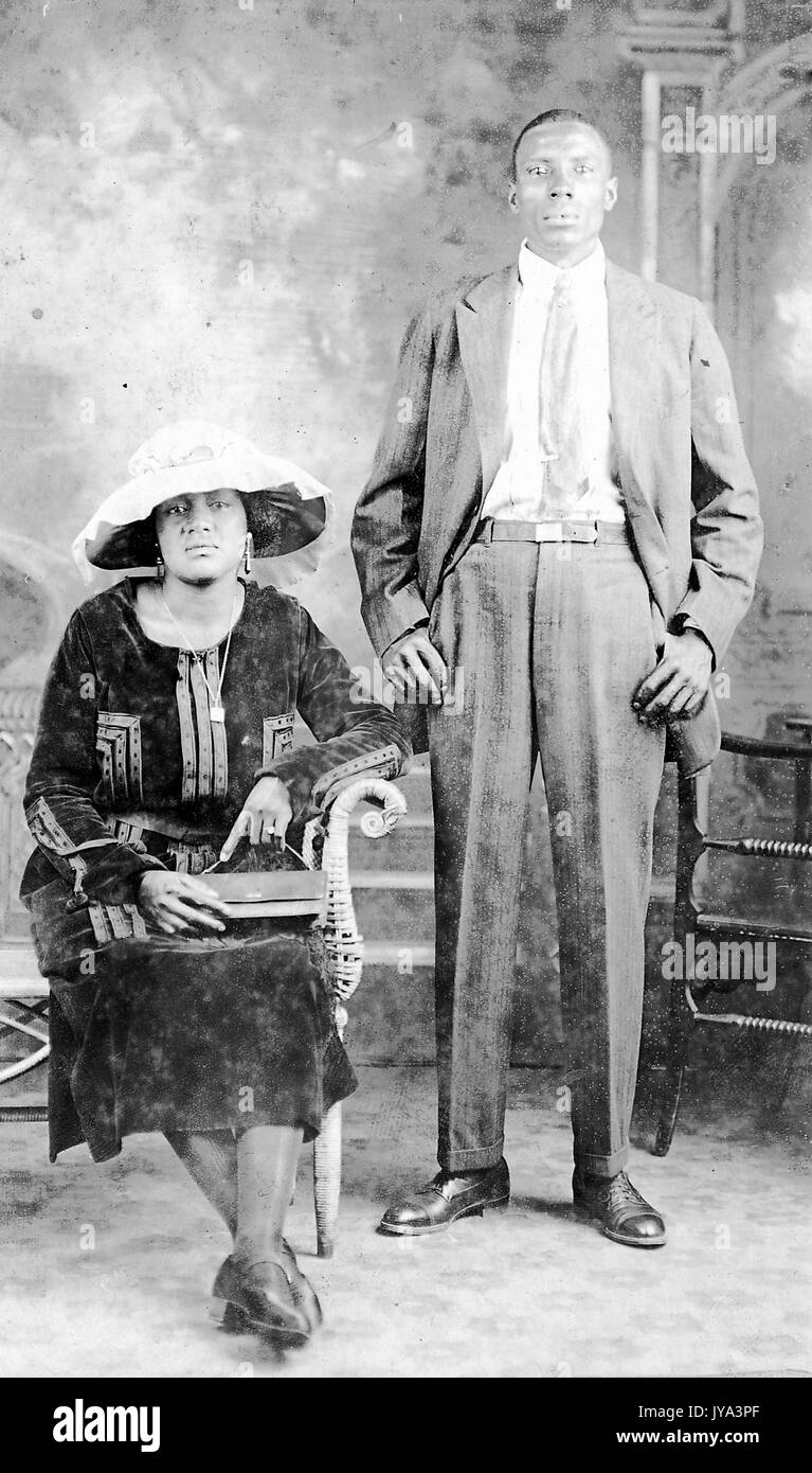 African American couple, the man standing with his hands on his hips and wearing suit, woman sitting on a bench and wearing a large brimmed hat, posing for photographs at a photographic studio, June 4, 1922. Stock Photo
