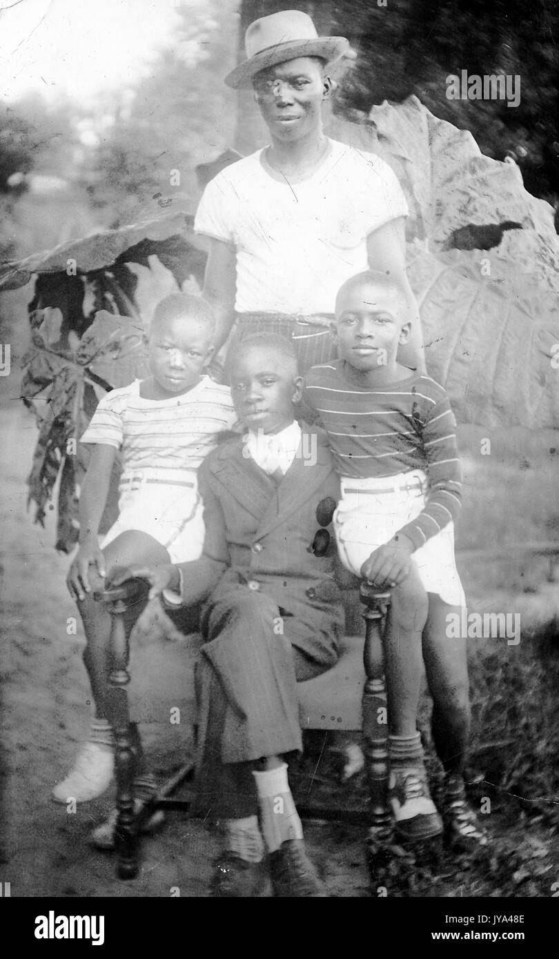 African-American family, a father posing for a portrait with his three sons, the father wearing a hat and standing, the sons sitting on a wooden chair, painted backdrop of trees, 1905. Stock Photo