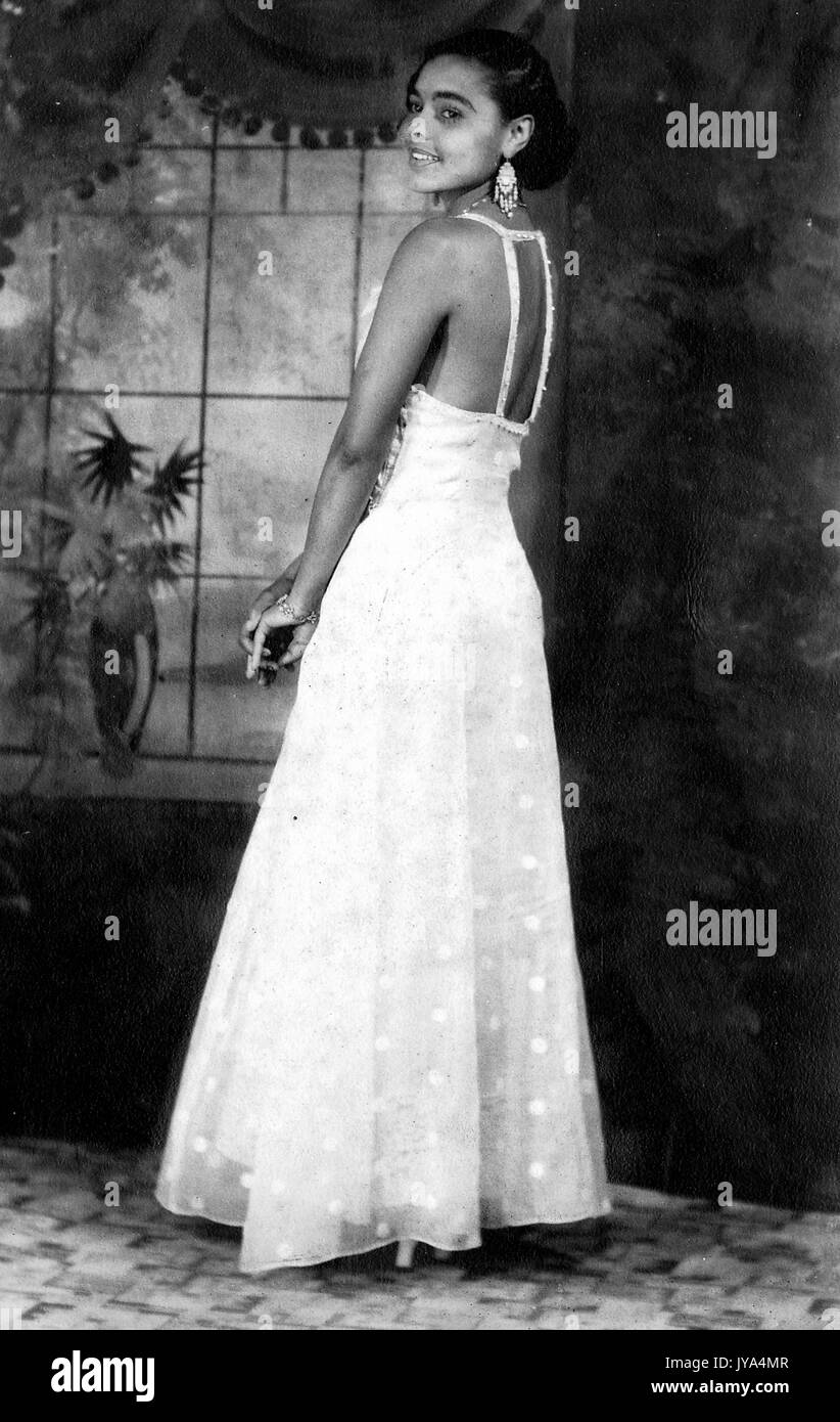 Pretty young African American girl posing for photographs in a white dress, view from behind looking over her shoulder at the camera and smiling, painted backdrop visible, 1932. Stock Photo