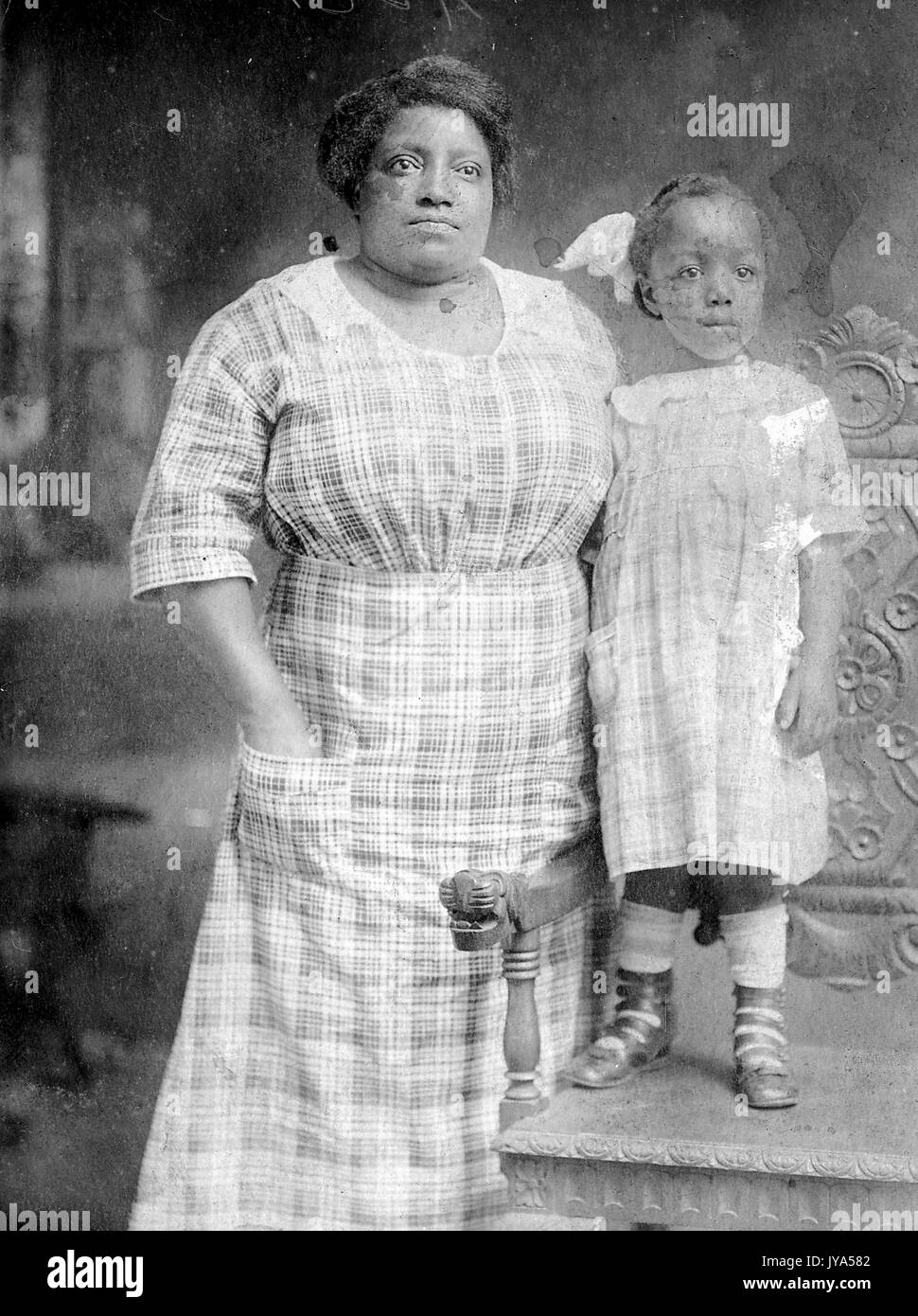 African-American mother and young girl posing for a studio portrait, the woman wearing a dress, the girl wearing a matching dress and standing on a chair, making her the same height as her mother, 1912. Stock Photo