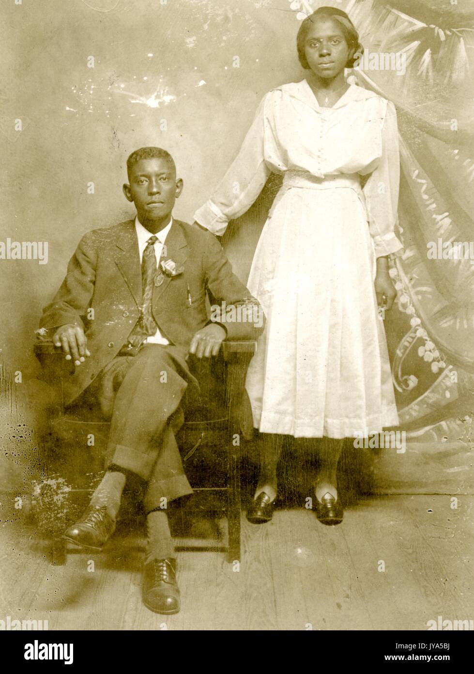 African-American couple, a man and a woman, posing for a studio portrait against a painted backdrop, the man sitting in a chair and wearing a suit with a flower in the pocket, the woman wearing a white dress, standing and resting her hand on the chair, both with neutral facial expressions, 1905. Stock Photo