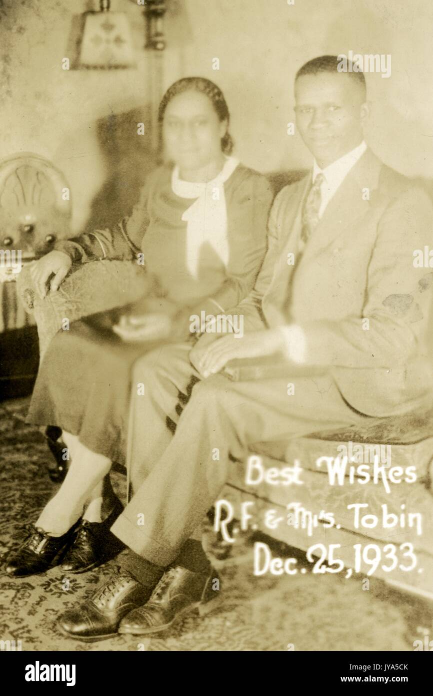 African-American couple seated on a couch in their home, posing for a portrait photo, the man wearing a suit and tie, the woman wearing a dress, both with their feet crossed and smiling slightly, with text reading Best Wishes RF and Mrs Tobin, on Christmas Day, some blurriness on the face of the woman, 1905. Stock Photo