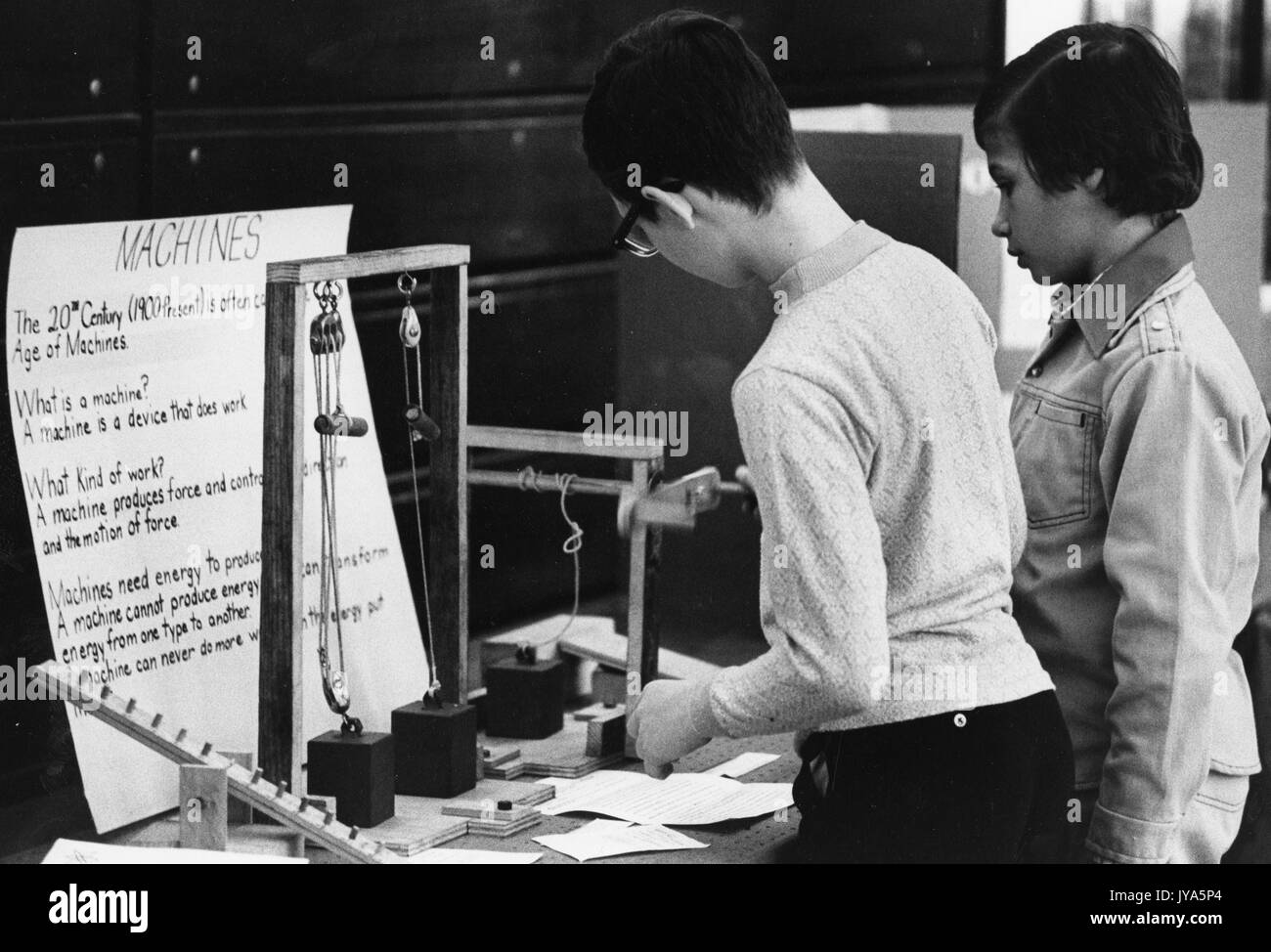 Two young male students investigate a project at a science fair about machines, interacting with a series of blocks and pulleys set up in front of a poster about machines in the 20th century. 1975. Stock Photo