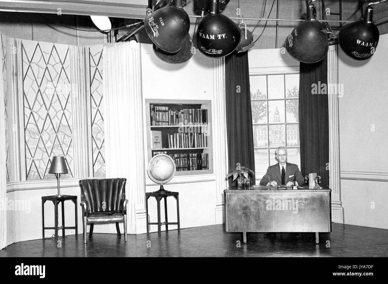 A view of one of the sets for the television show the Johns Hopkins Science Review, host Lynn Poole is sitting alone at a desk in front of one of two large windows, stage lights hang above him, 1955. Stock Photo