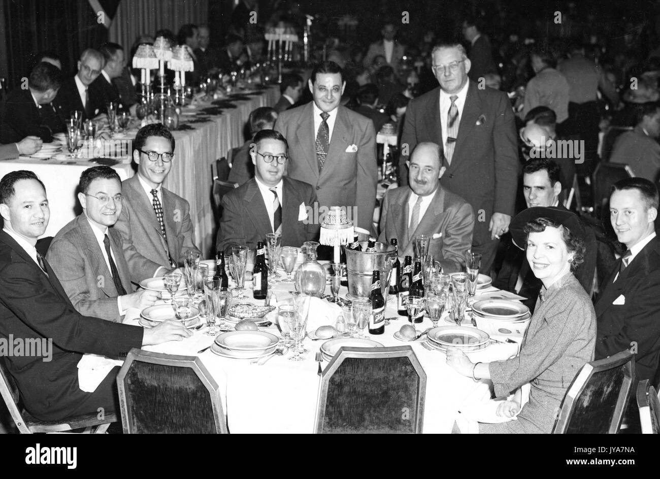 Herb Cahan, Henry Fischer, Joel Chaseman, Jack Harrington, Norman Kal, 'Mac', Ken Carten, Gray, and Tony are all dining and sitting around a circular table at an event to honor participants in the television show The Johns Hopkins Science Review, the table is covered in silverware, dishes, and beer bottles, there are many more tables behind them, 1955. Stock Photo