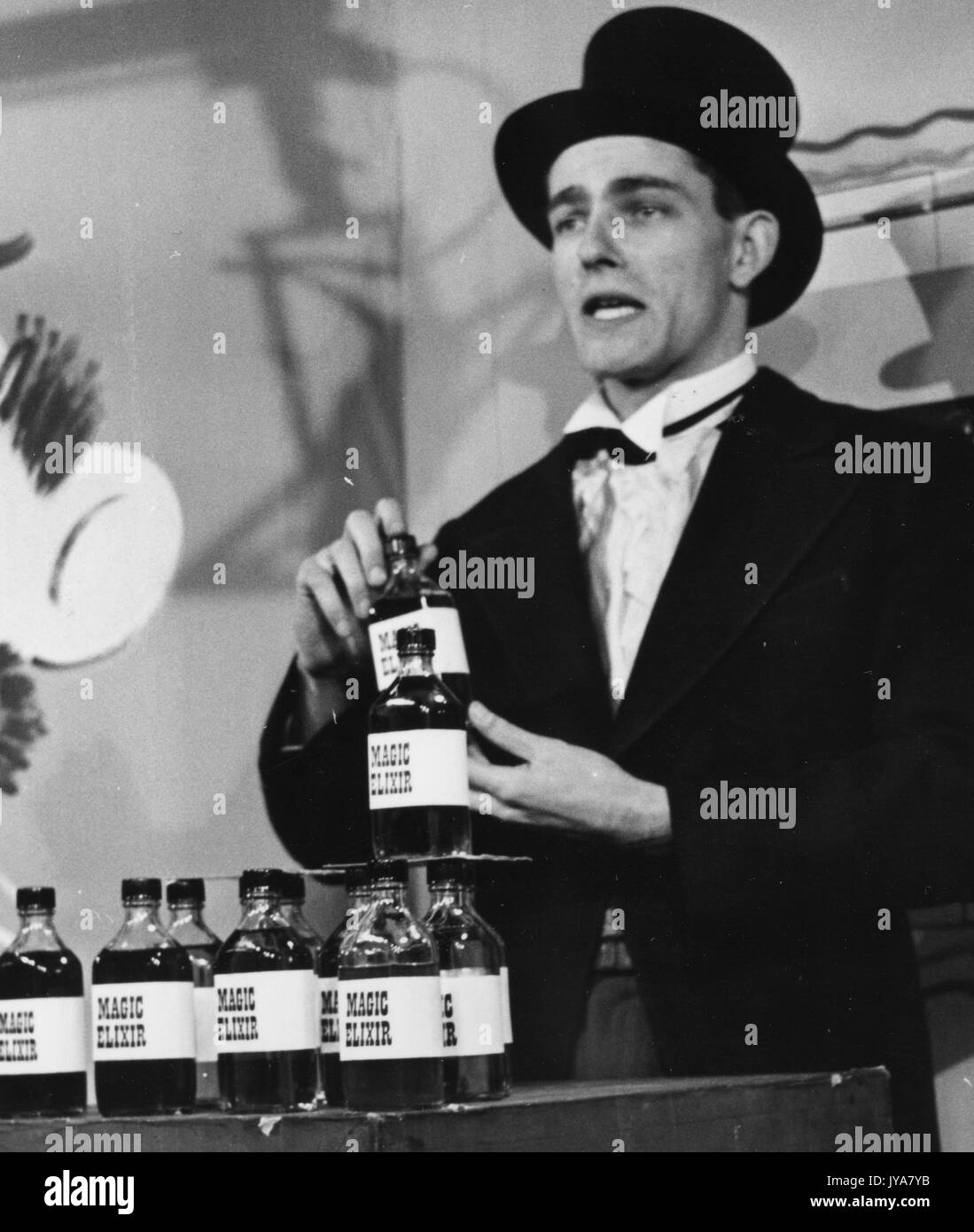 Actor Edmond Levy is on set filming for The Johns Hopkins Science Review, he is wearing a suit, a bow tie, and a black top hat, he is holding a bottle labeled Magic Elixir, he is standing behind a table holding many bottles labeled Magic Elixir, 1955. Stock Photo