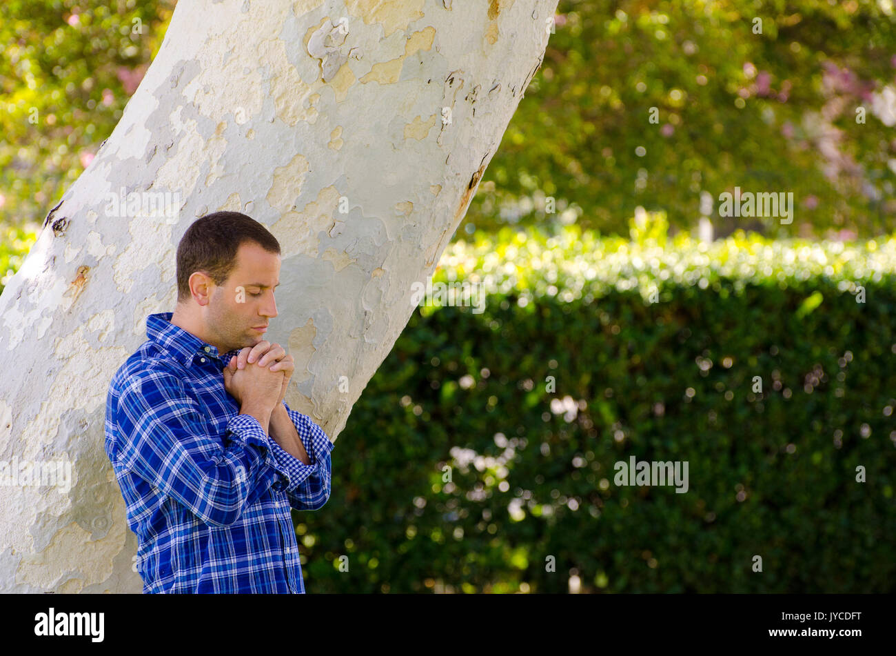 Man praying alone outside by a tree trunk. Stock Photo
