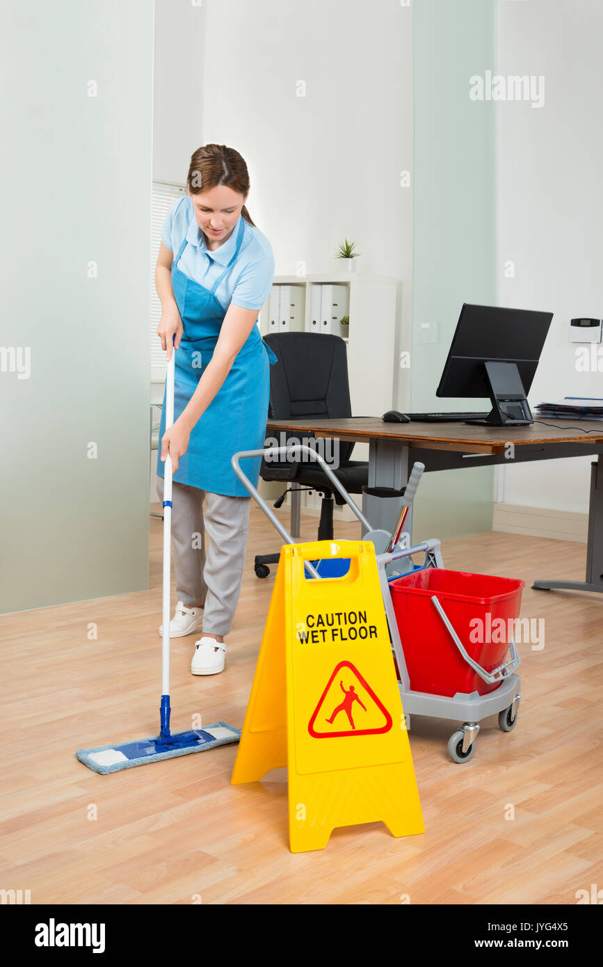 Happy Female Janitor With Cleaning Equipments Cleaning Hardwood Floor In Office Stock Photo