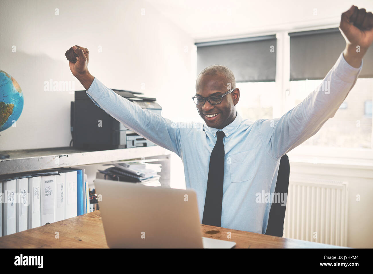 Smiling African businessman celebrating with his arms raised in the air while sitting at a desk in his home office working on a laptop Stock Photo