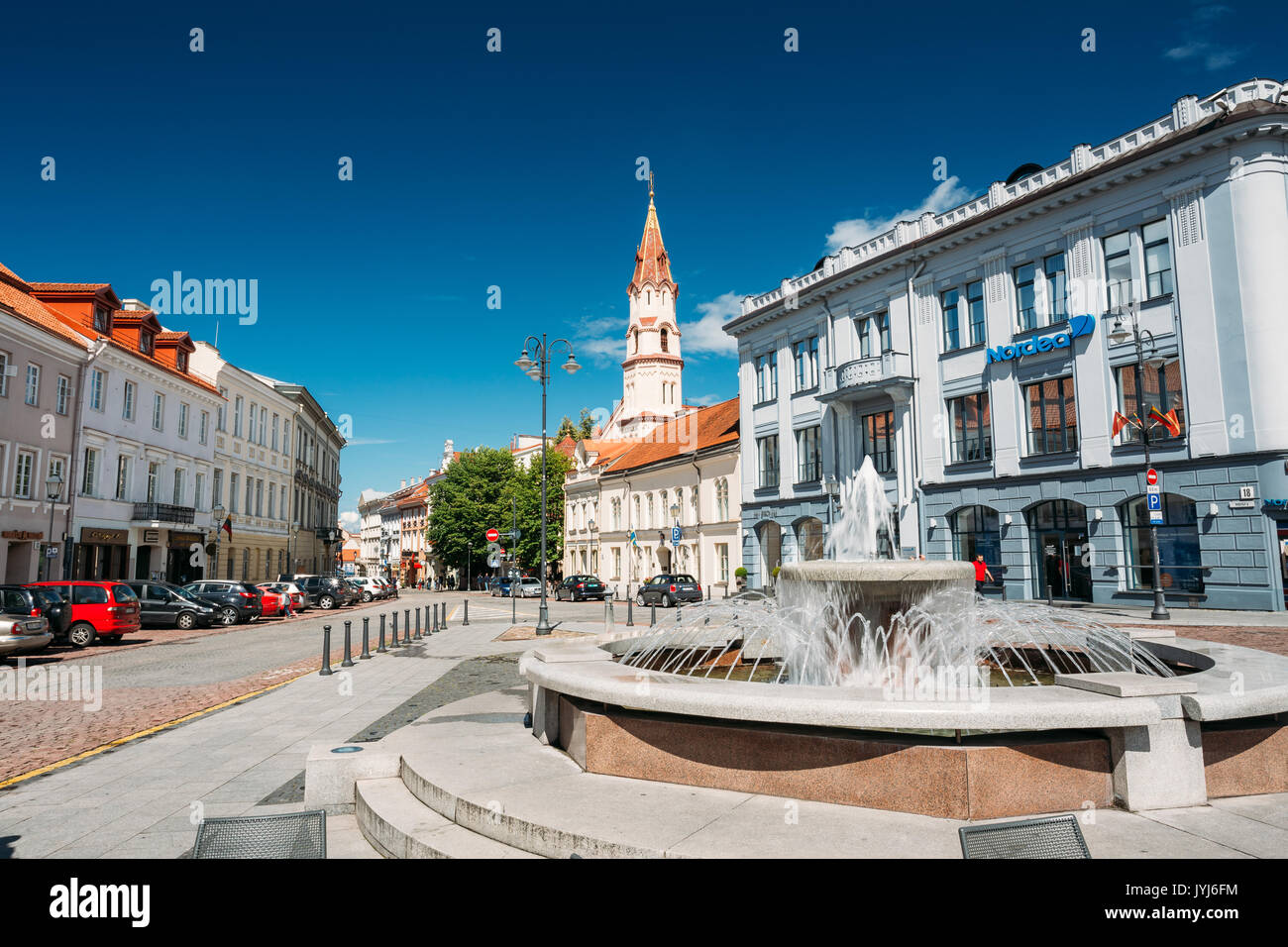 Vilnius, Lithuania - July 5, 2016: Town Hall Square Fountain In Rotuses Square In Old Town. St. Nicholas Church In Sunny Summer Day. Popular Touristic Stock Photo