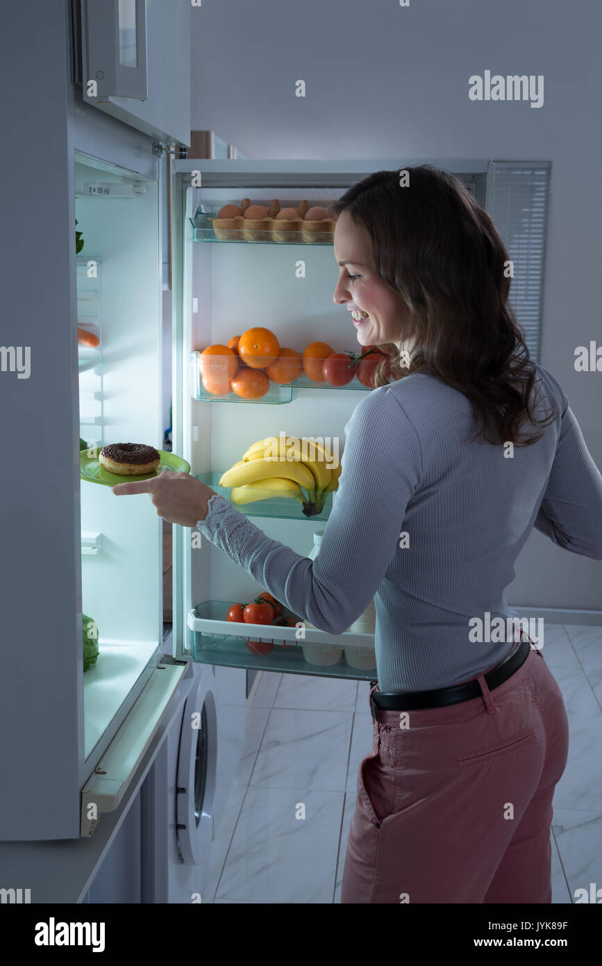 Happy Young Woman Taking Donut From Refrigerator At Night Stock Photo