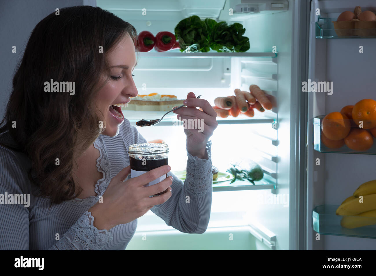 Young Woman Eating In Front Of Fridge In Kitchen Stock Photo
