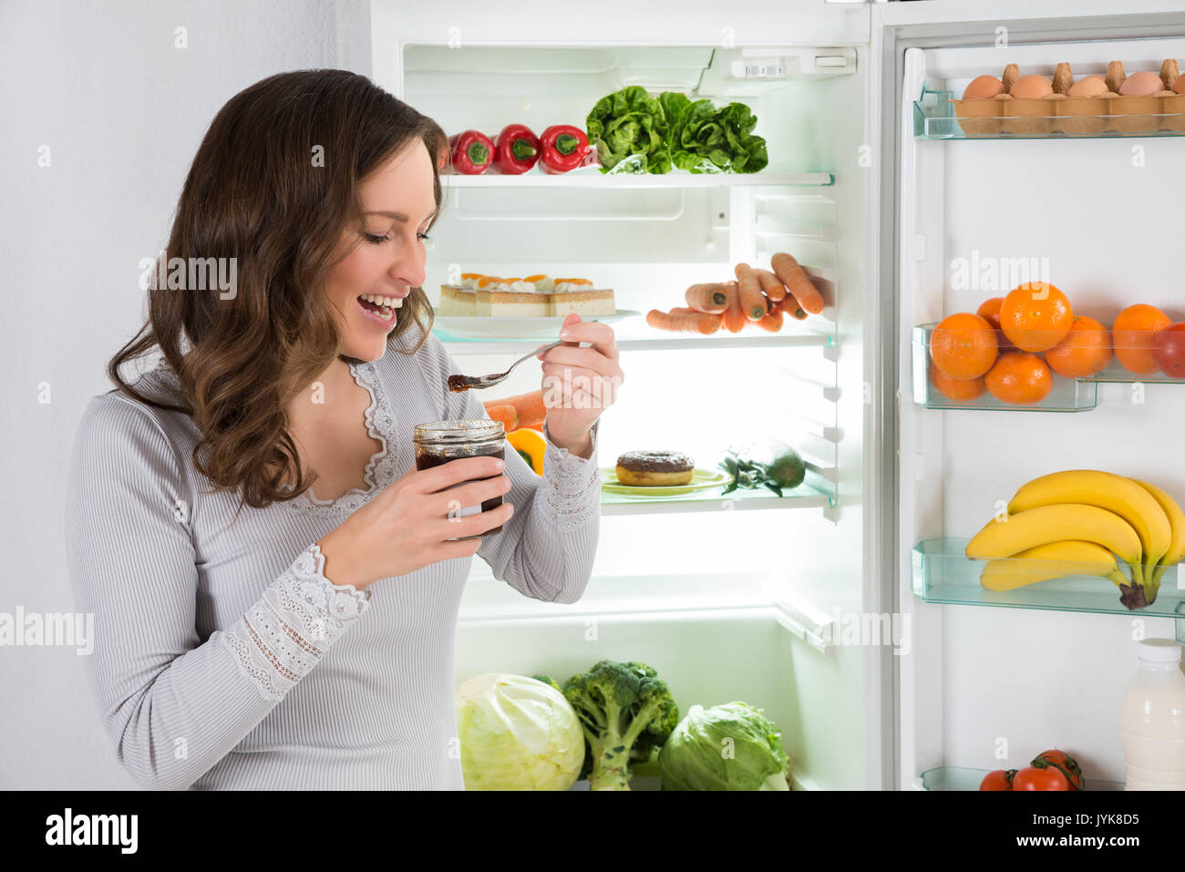Young Woman Eating In Front Of Fridge In Kitchen Stock Photo