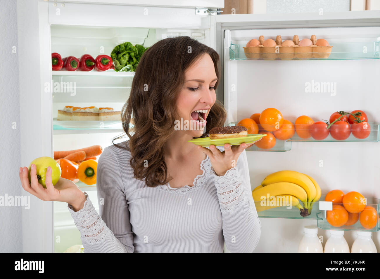 Young Woman With Green Apple Eating Donut In Front Of Fridge Stock Photo