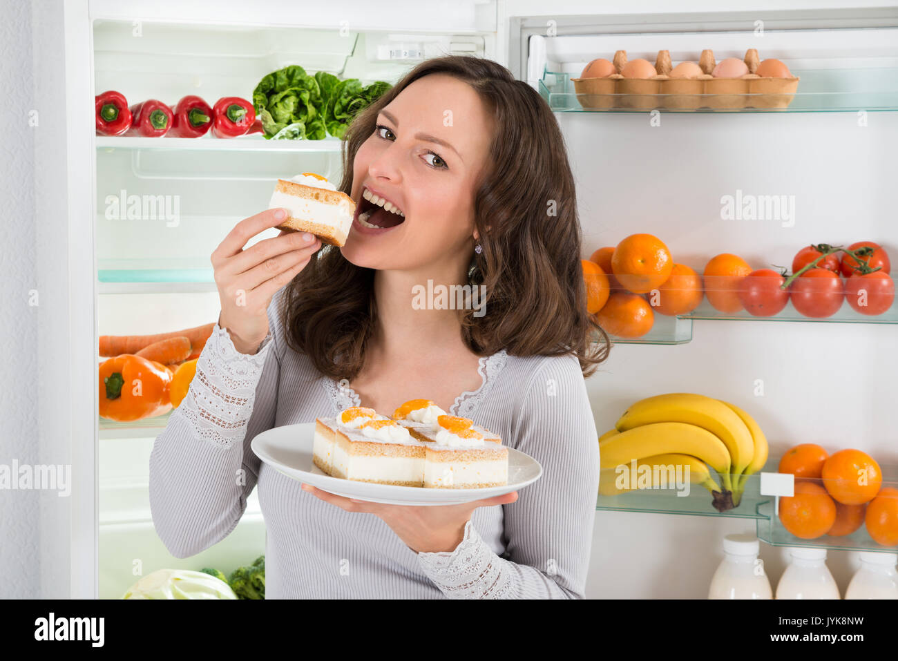 Young Woman Eating Slice Of Cake Near The Open Refrigerator Stock Photo