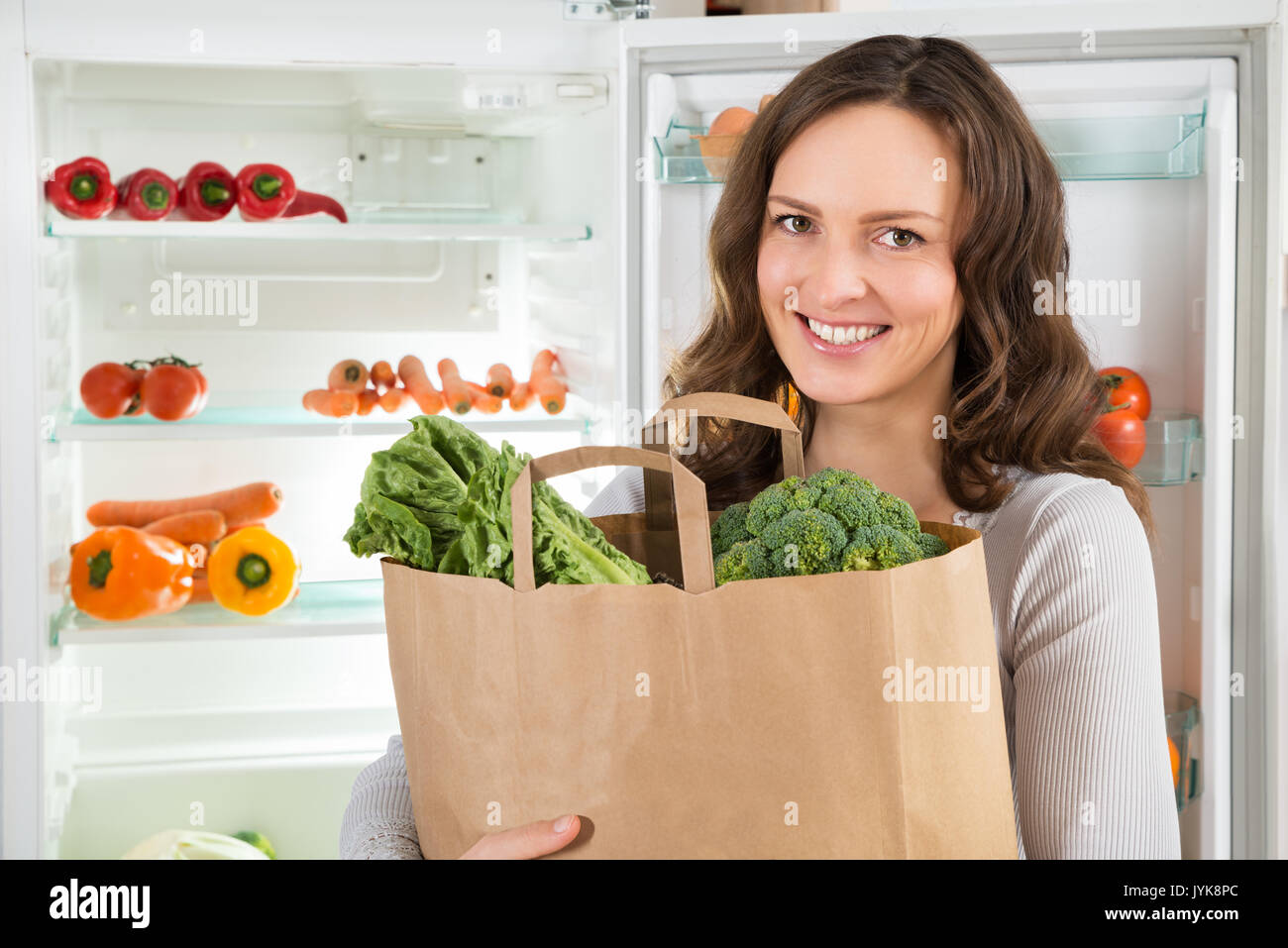 Happy Woman Holding Grocery Shopping Bag With Vegetables In Front Of Open Refrigerator Stock Photo