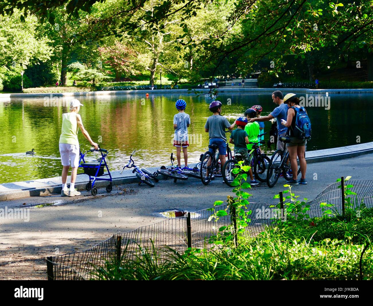 Elderly woman with walker observes family with bicycles - mother, father, five boys - stopped next to a pond in Central Park, New York, NY, USA. Stock Photo