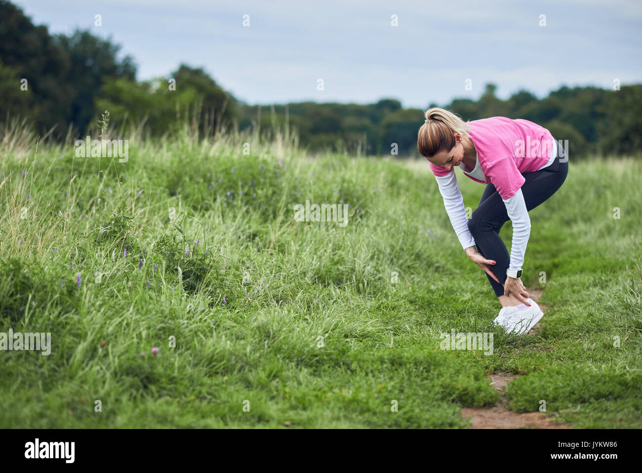 An exercising woman in fit wear while running outdoors, stop to clutch an injured ankle. Stock Photo