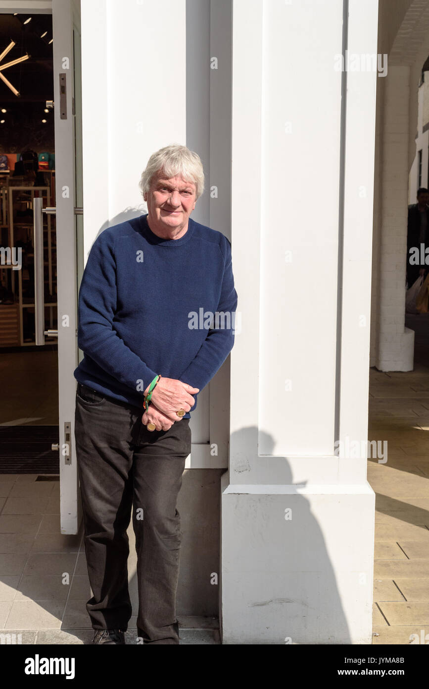 White haired man in casual clothing with hands held together leans against a white painted wall waiting utside a shop Stock Photo