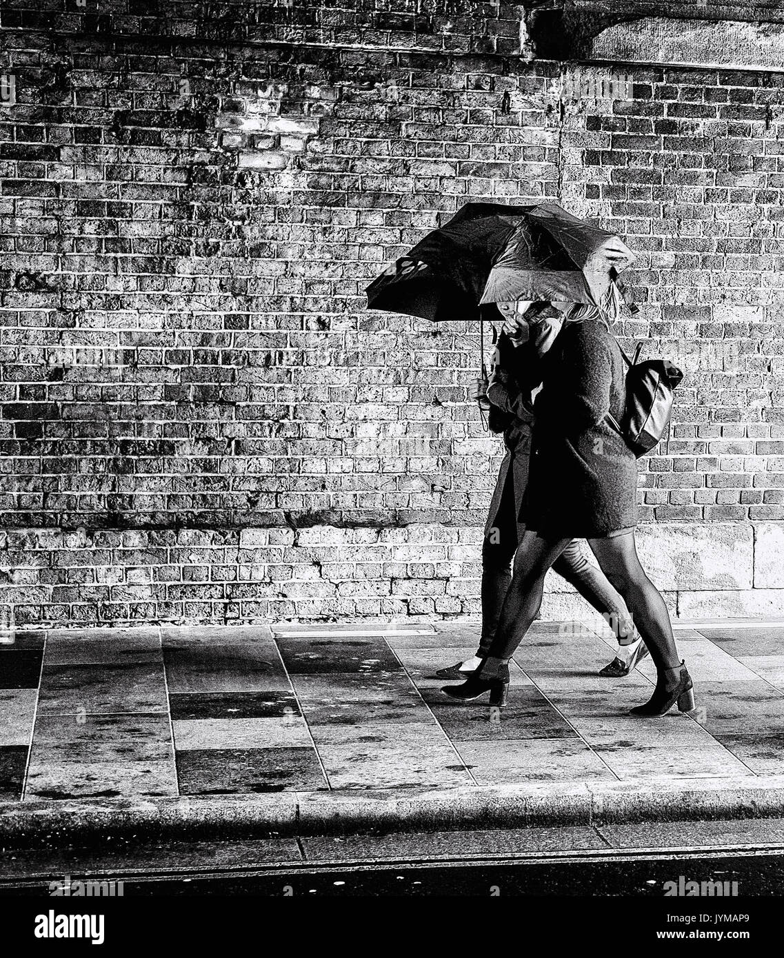 Two business women in smart clothing hold and umbrella as they walk under an old concrete bridge Stock Photo