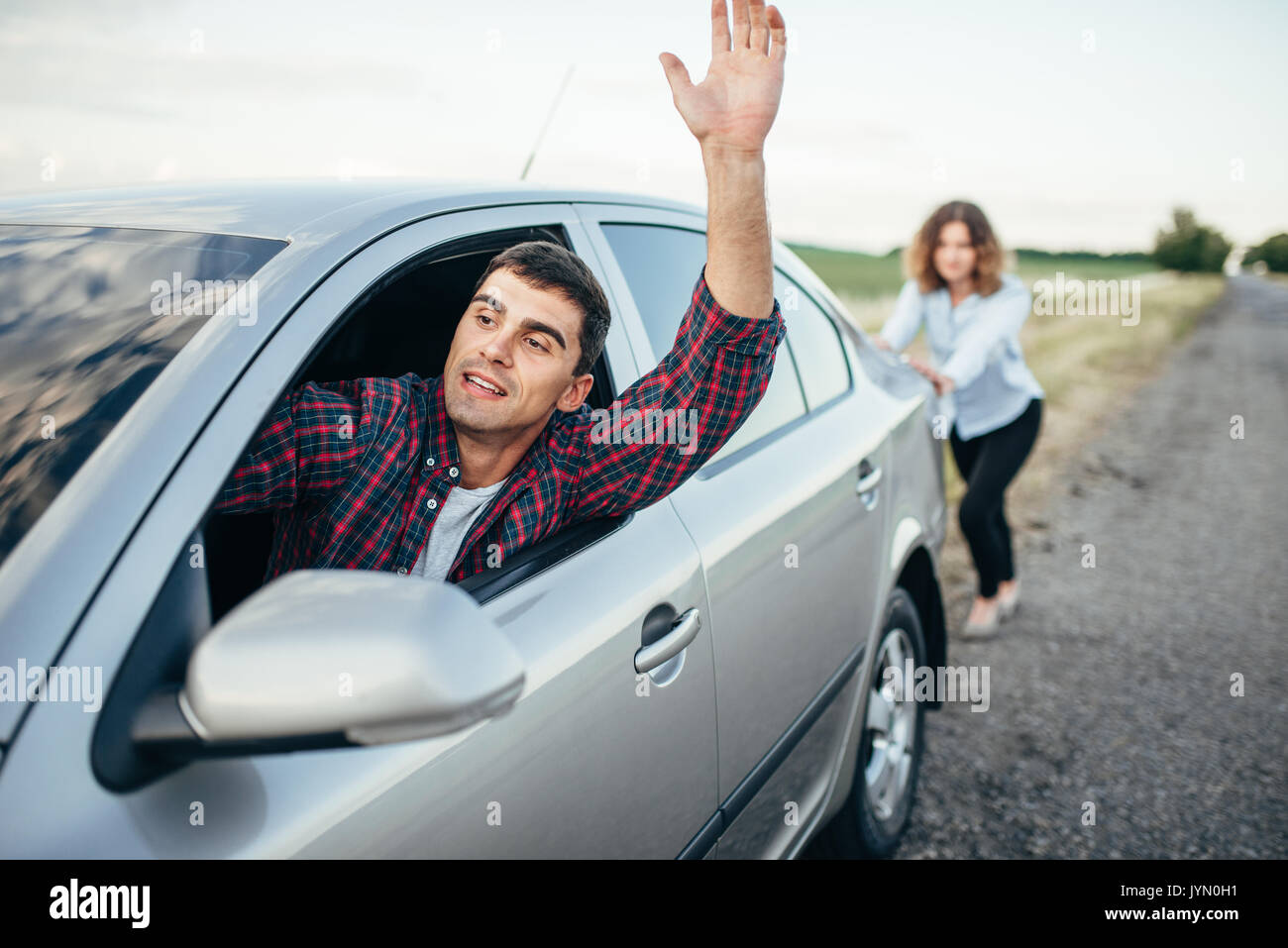 Woman pushing a broken car down the road, smiling man driver. Vehicle with trouble Stock Photo