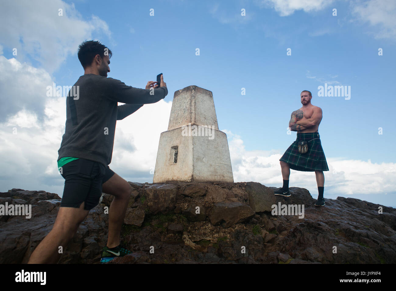 Photo-opportunity on the peak. A muscular topless man in a kilt is photographed by his friend on the top of the peak.  The peak of King Arthur's Seat  Stock Photo