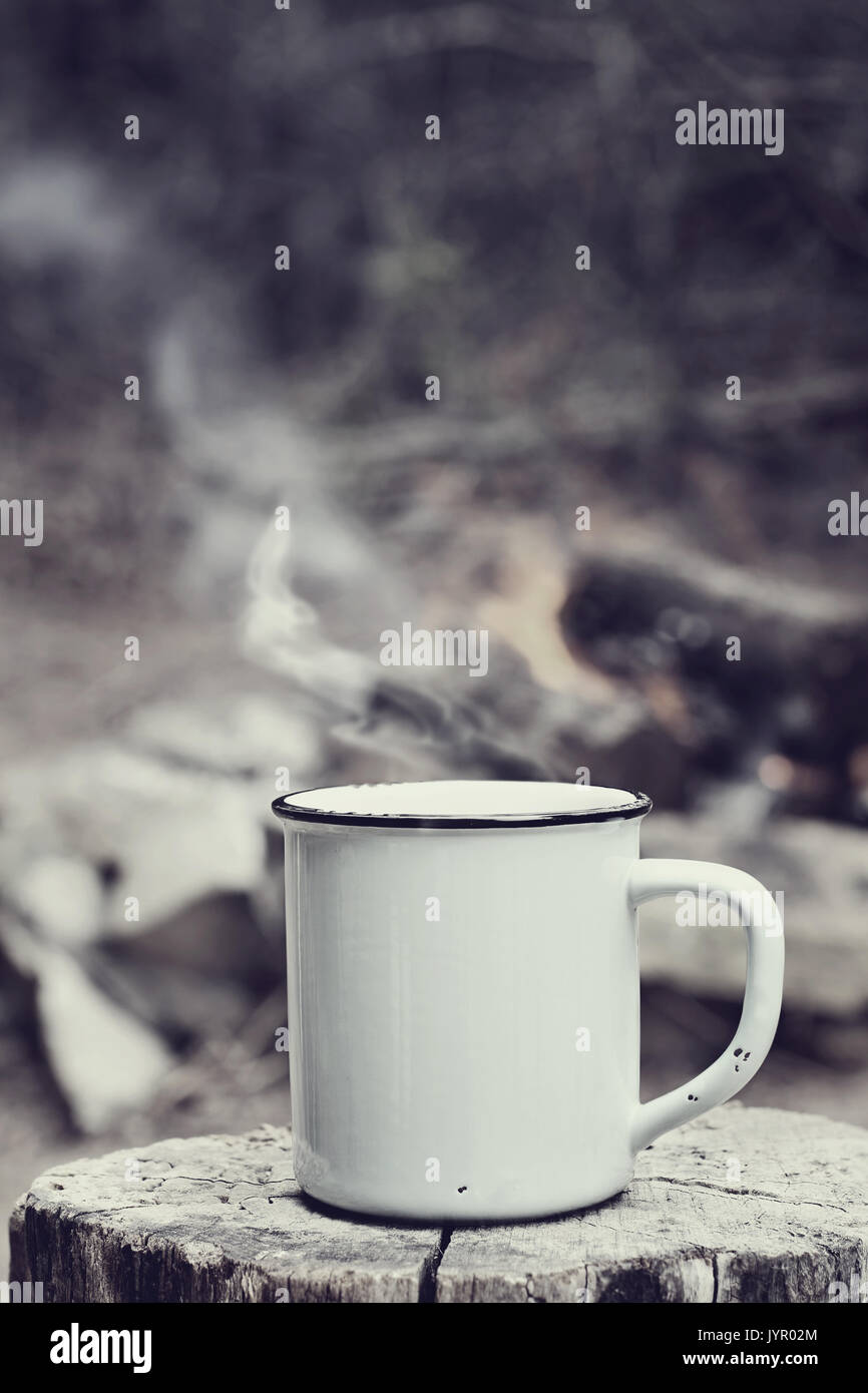 Cup of hot steaming coffee sitting on an old log by an outdoor campfire with a vintage antique edit. Extreme shallow depth of field with selective foc Stock Photo