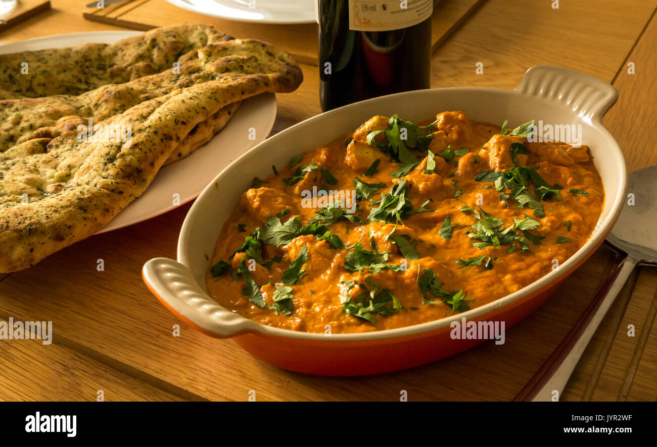 Close up of serving dish of chicken curry with naan bread and bottle of red wine on oak table Stock Photo