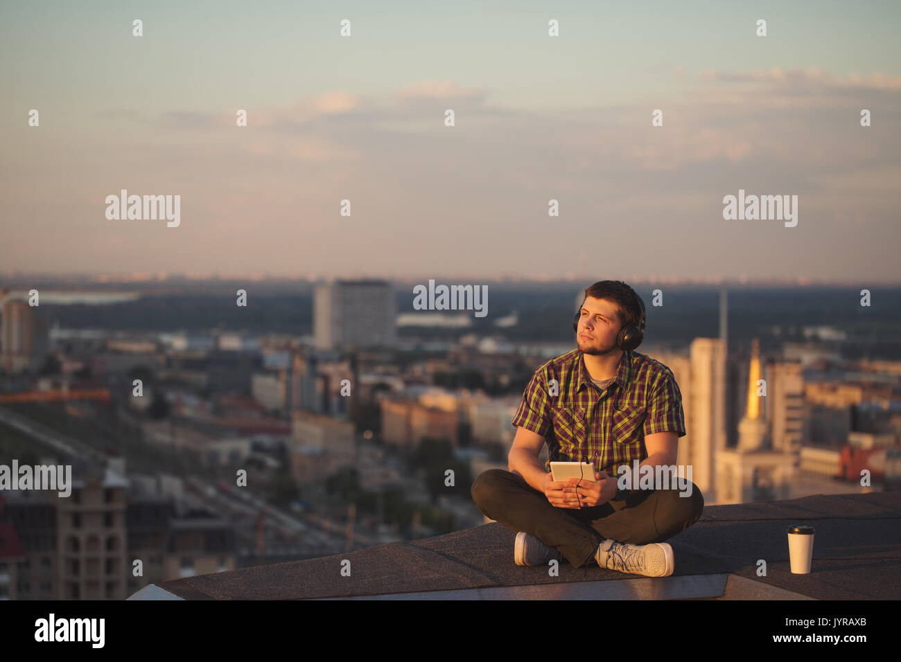 A hipster guy listens to music on headphones on the roof of a high-rise building. He looks into the distance and enjoys the moment. Freelancer in sear Stock Photo