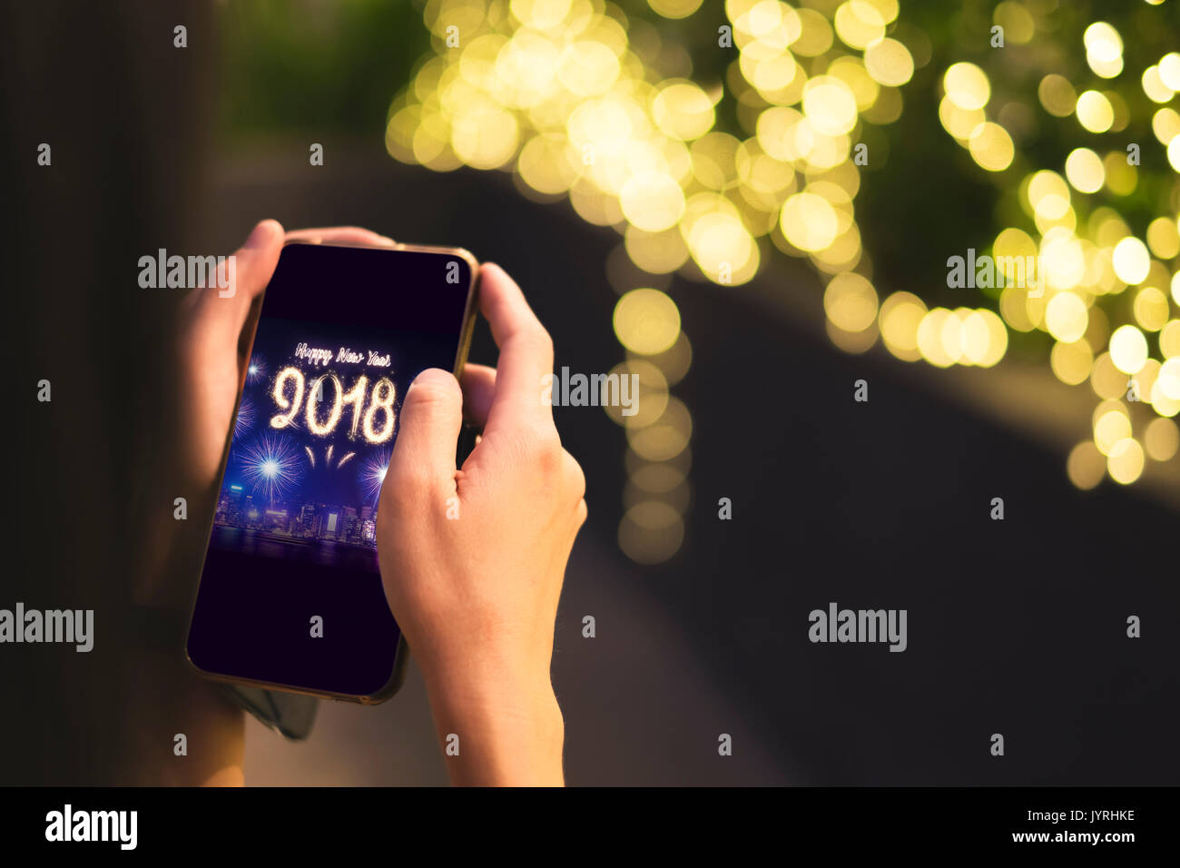 Woman Hand holding mobile with Happy new year 2018 fireworks over cityscape on screen with light bokeh background,Digital greeting card holiday. Stock Photo