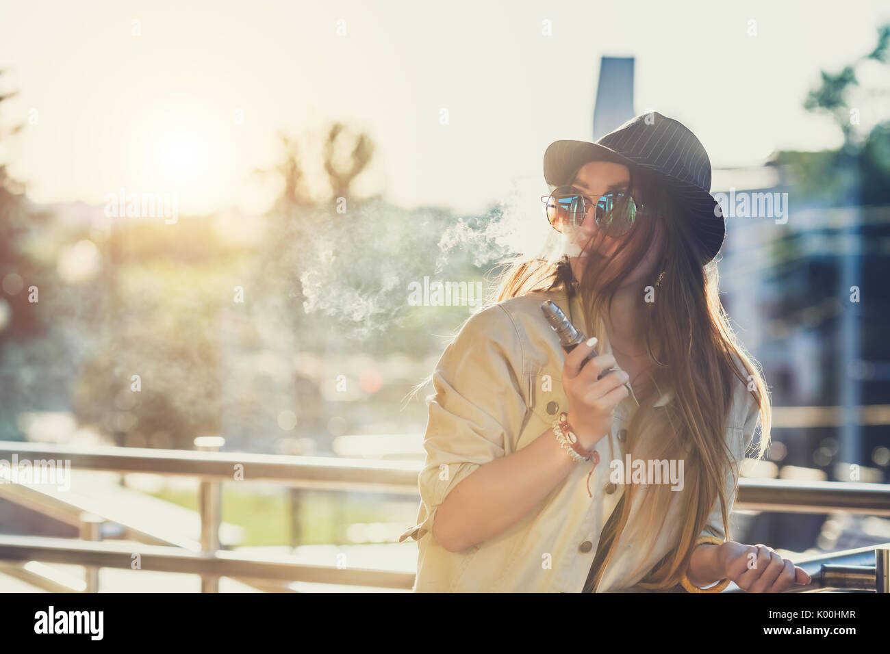 Pretty young hipster woman in black hat vape ecig, vaping device at the sunset. Toned image. Stock Photo