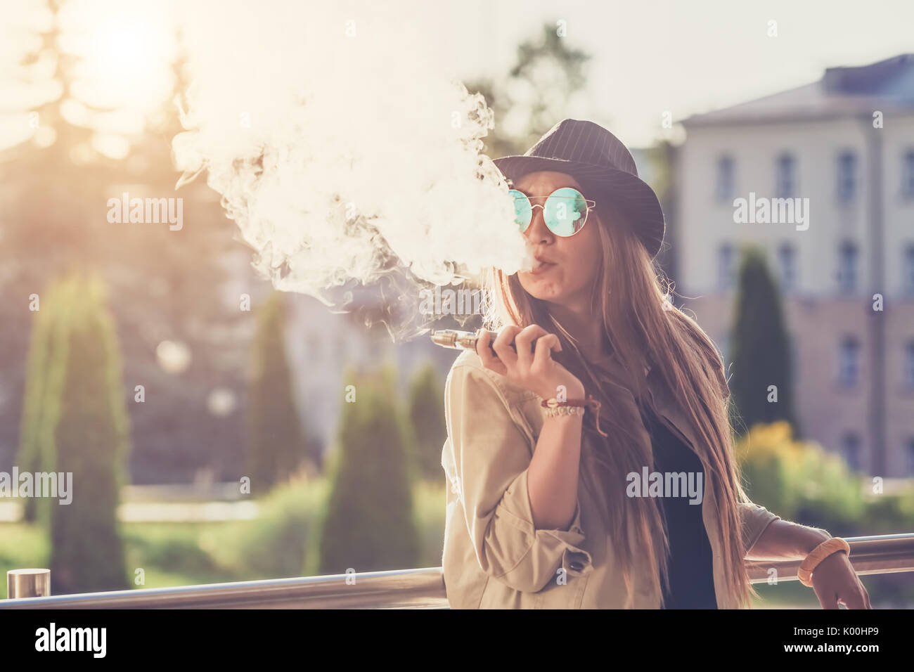 Pretty young hipster woman in black hat vape ecig, vaping device at the sunset. Toned image. Stock Photo