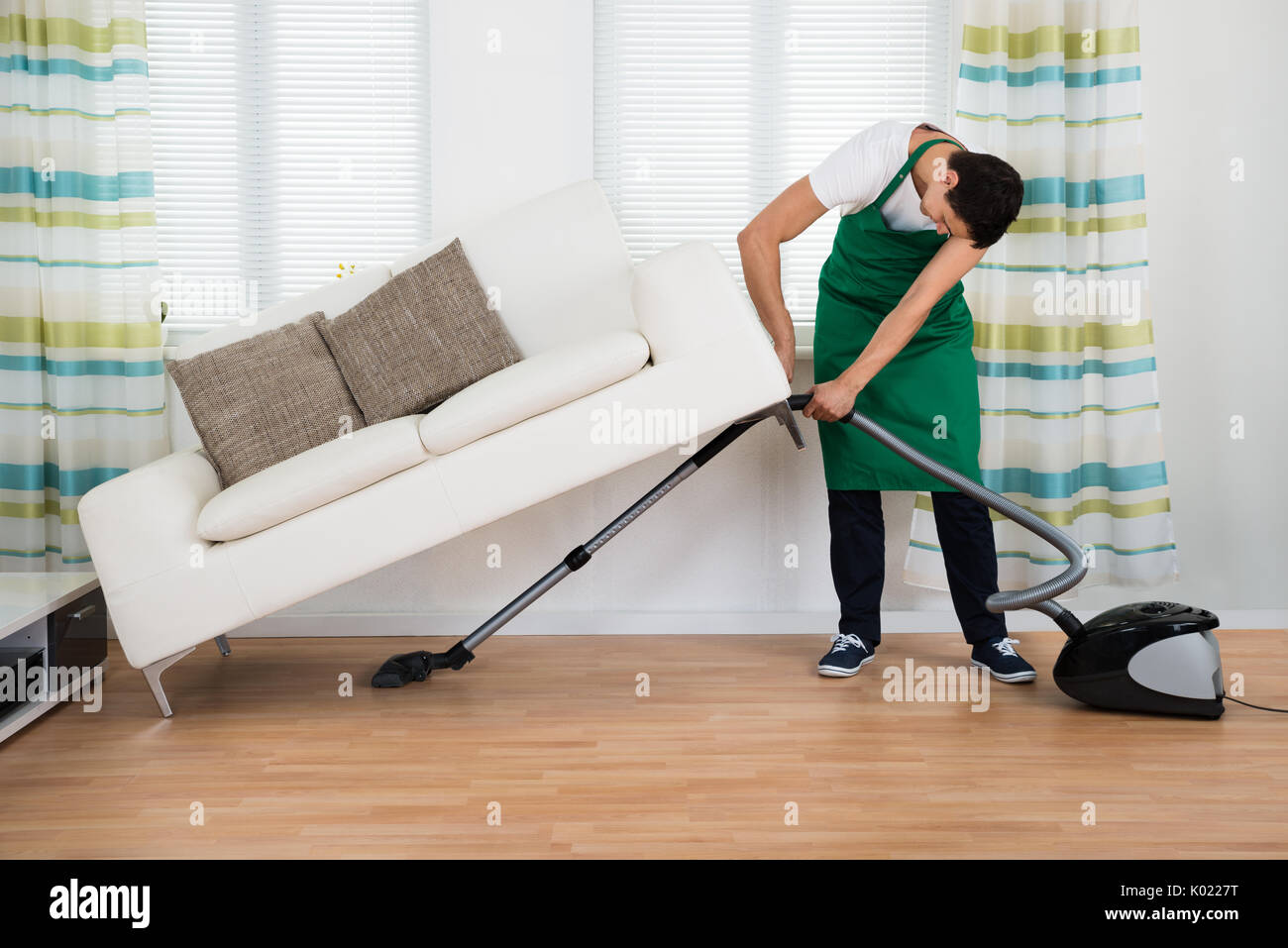 Full length of man lifting couch while cleaning hardwood floor with vacuum cleaner at home Stock Photo