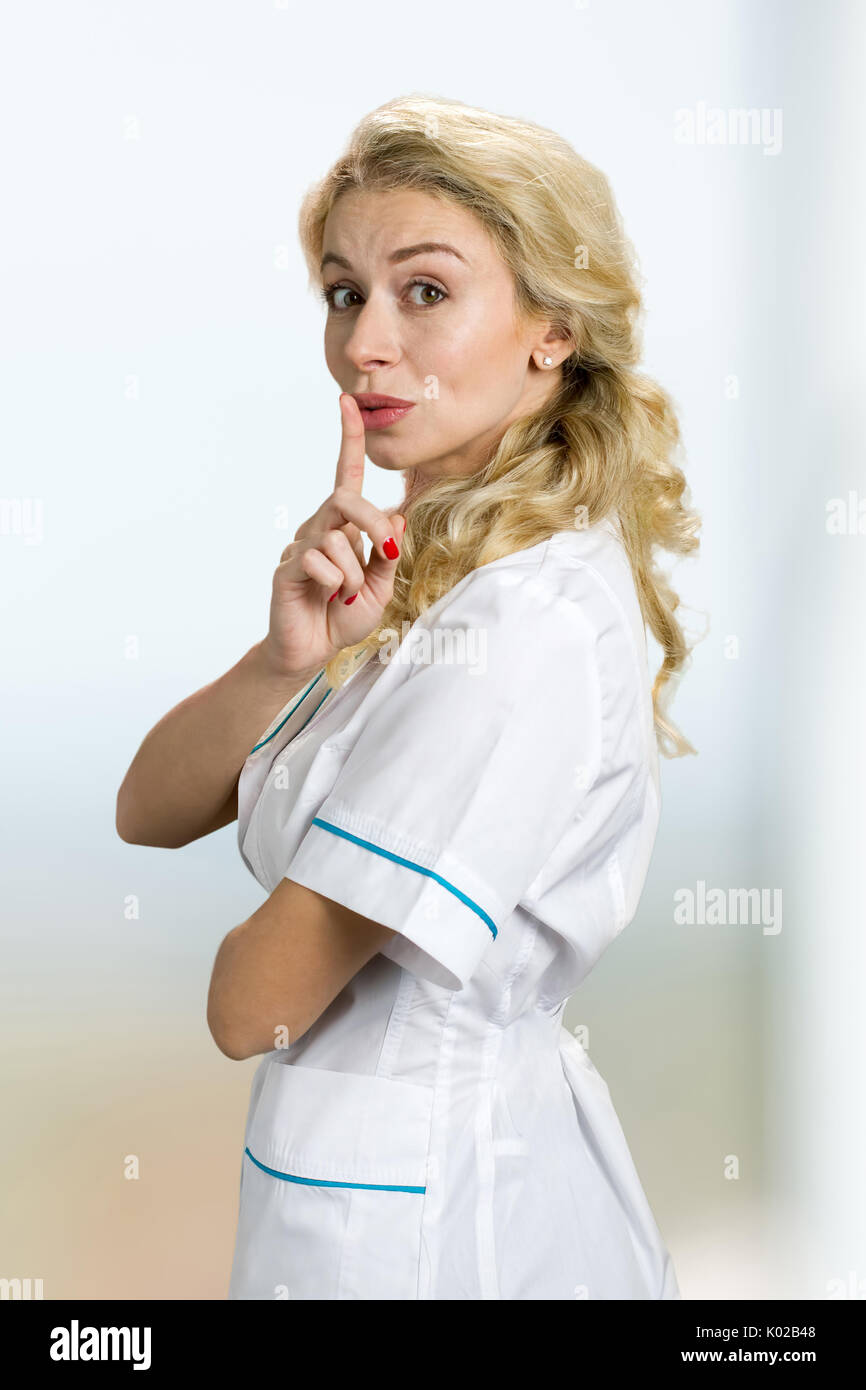 Attractive blond nurse gesturing silence. Young woman in lab coat making silence gesture, shhh. Keep the silence gesture. Stock Photo