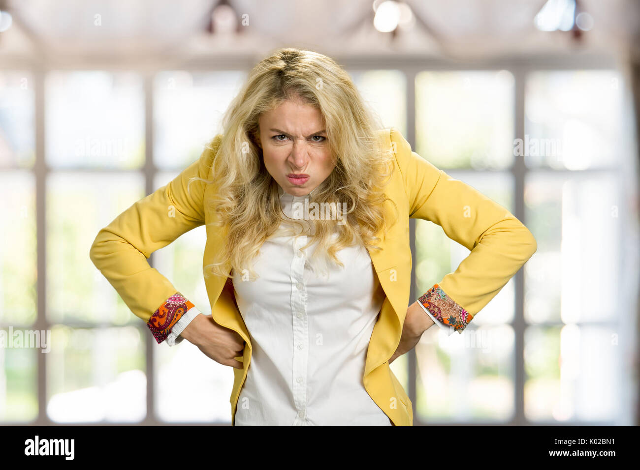 Aggressive frowning young blonde woman. Frustrated european woman in formal wear holding hands on hips with negative emotions, blurred background. Stock Photo