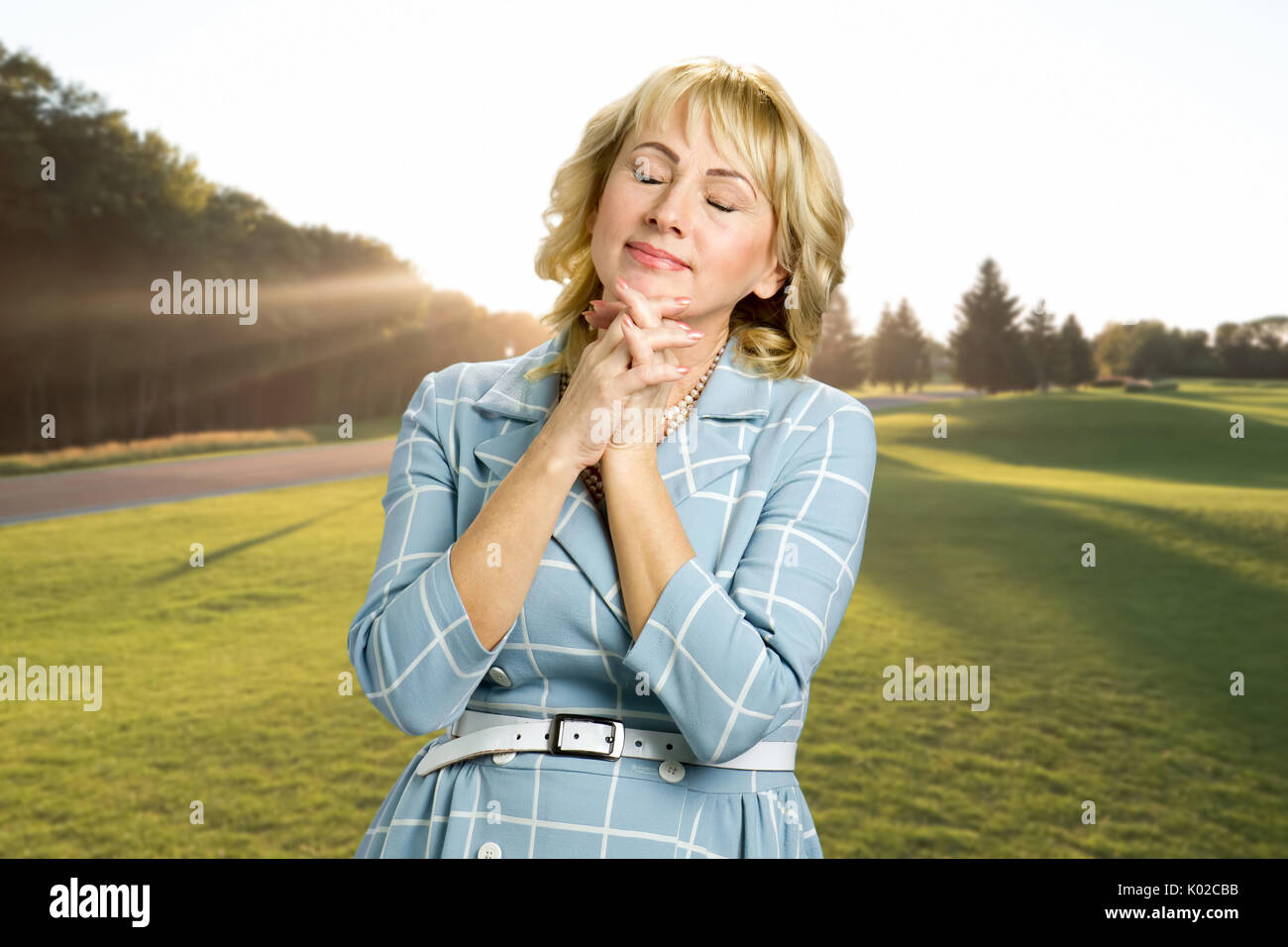 Relaxing mature woman outdoor. Middle aged woman with clasping hands under chin and closed eyes on nature background. Stock Photo