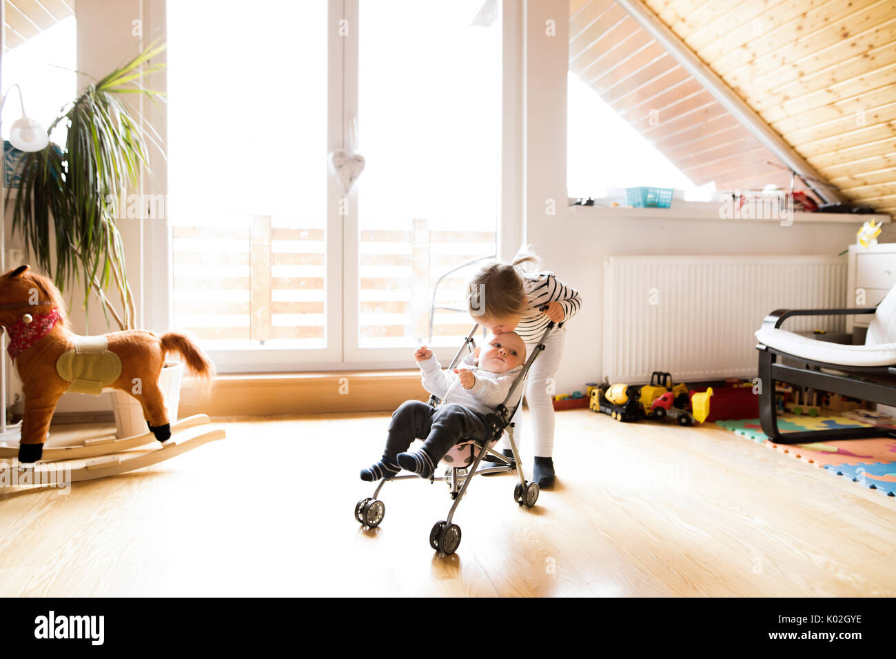 Little girl at home with baby brother in toy stroller. Stock Photo