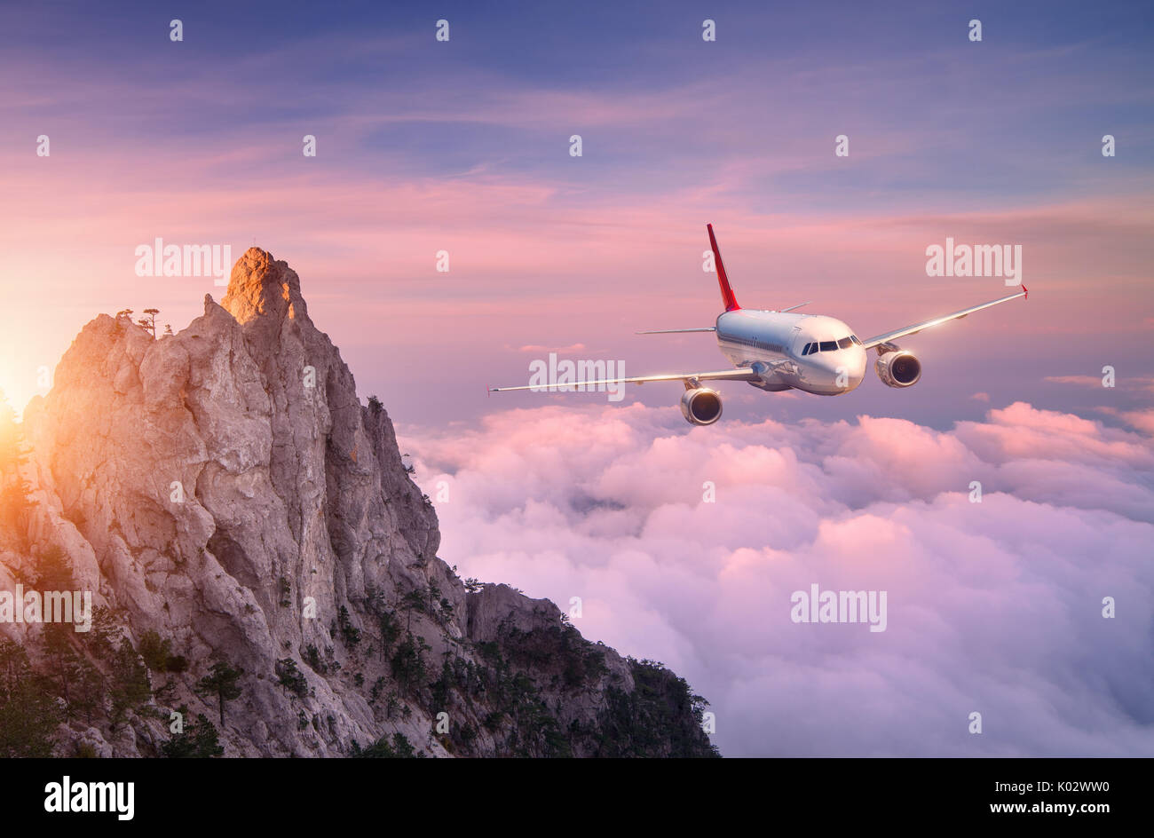 Airplane is flying over clouds at sunset. Landscape with white passenger airplane, rocks, sea and purple sky with sun in summer. Passenger aircraft is Stock Photo