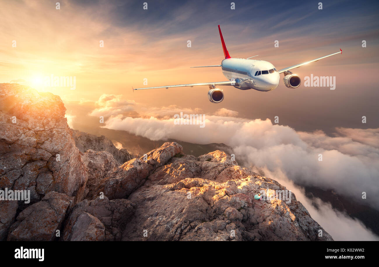 Airplane is flying over clouds at sunset. Landscape with white passenger airplane, mountains, sea and orange sky with sun in summer. Passenger aircraf Stock Photo