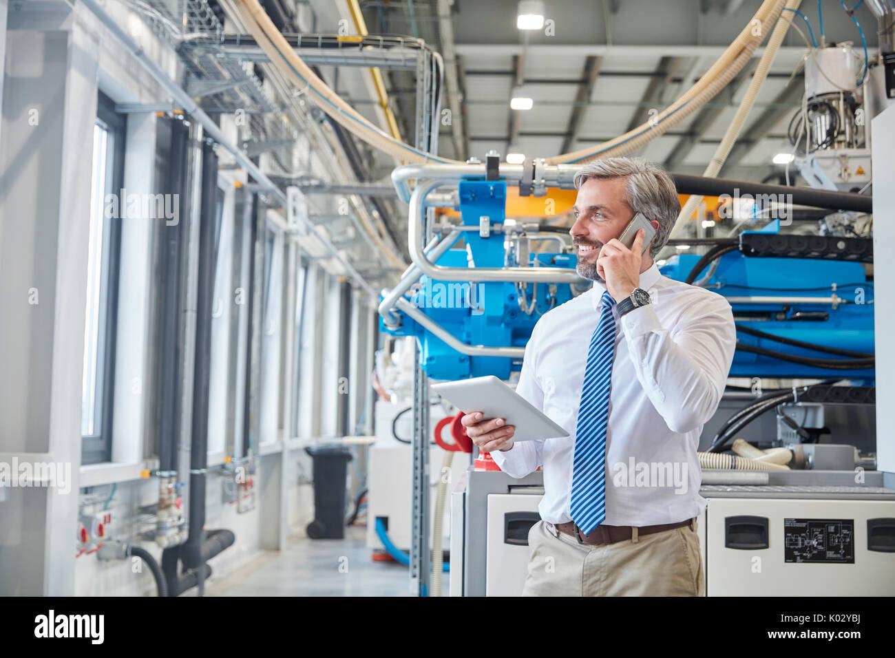 Male supervisor with digital tablet talking on cell phone next to plastic industrial molder machinery in factory Stock Photo