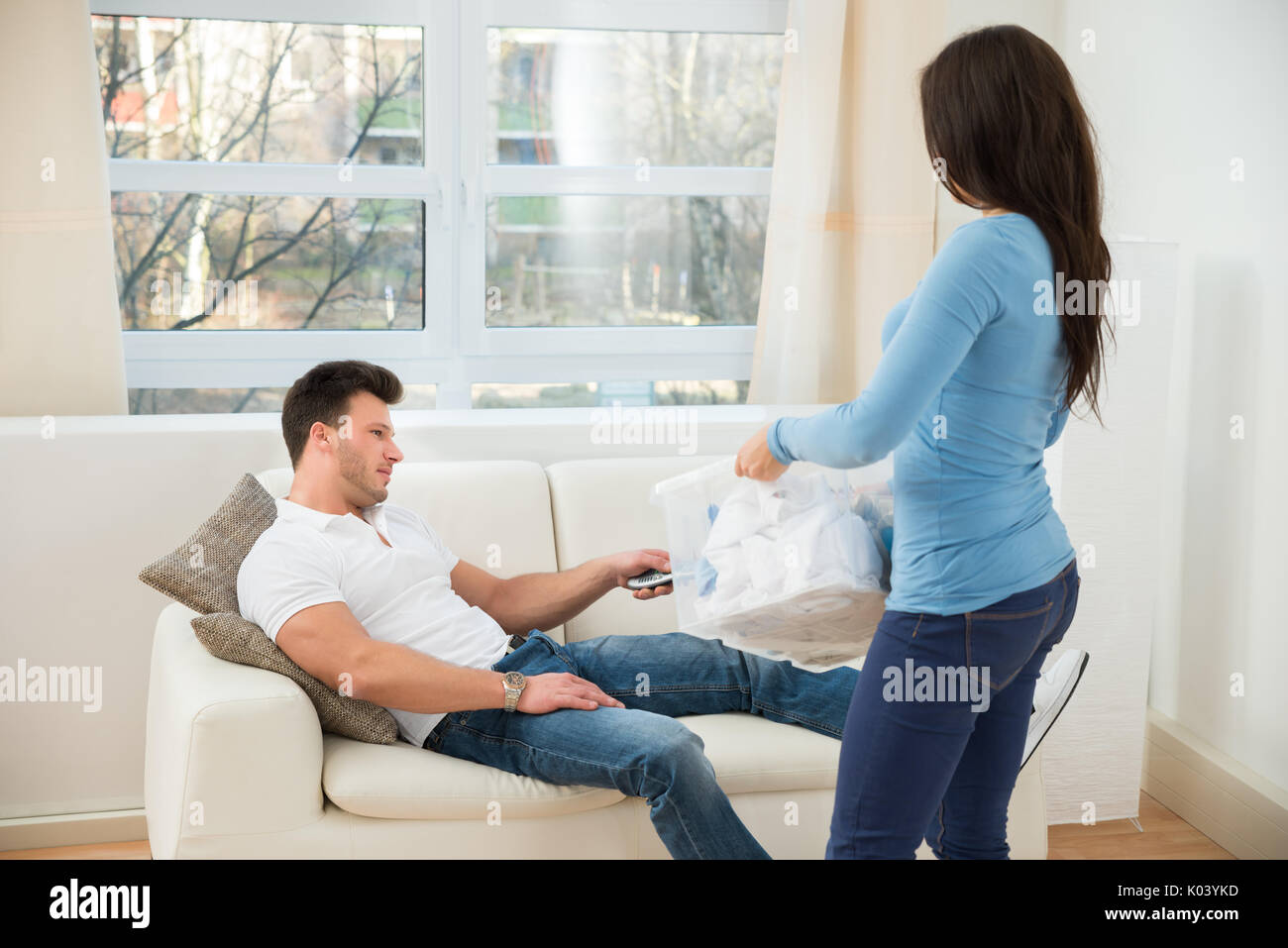 Woman Holding Laundry Basket Looking At Man Using Remote Control Stock Photo