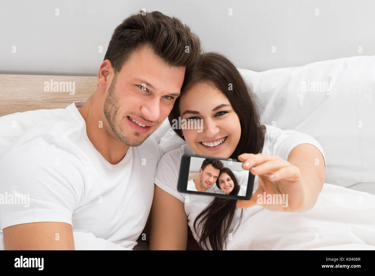 Happy Young Couple Lying On Bed Taking A Selfie With Mobile Phone Stock Photo