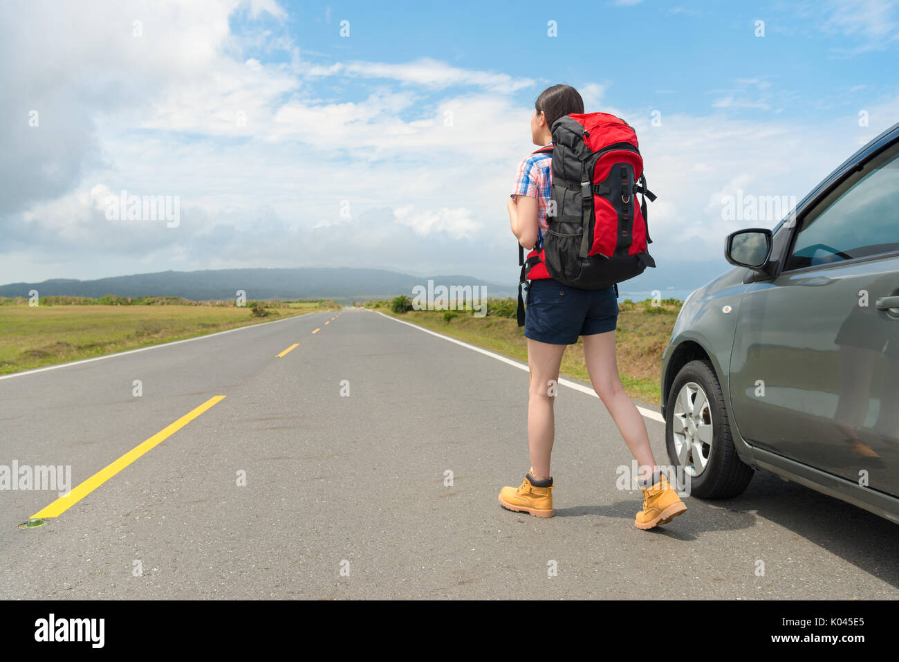 elegant leisurely female backpacker standing on asphalt road watching distant mountain scenery and parked car in roadside at summer vacation travel. Stock Photo