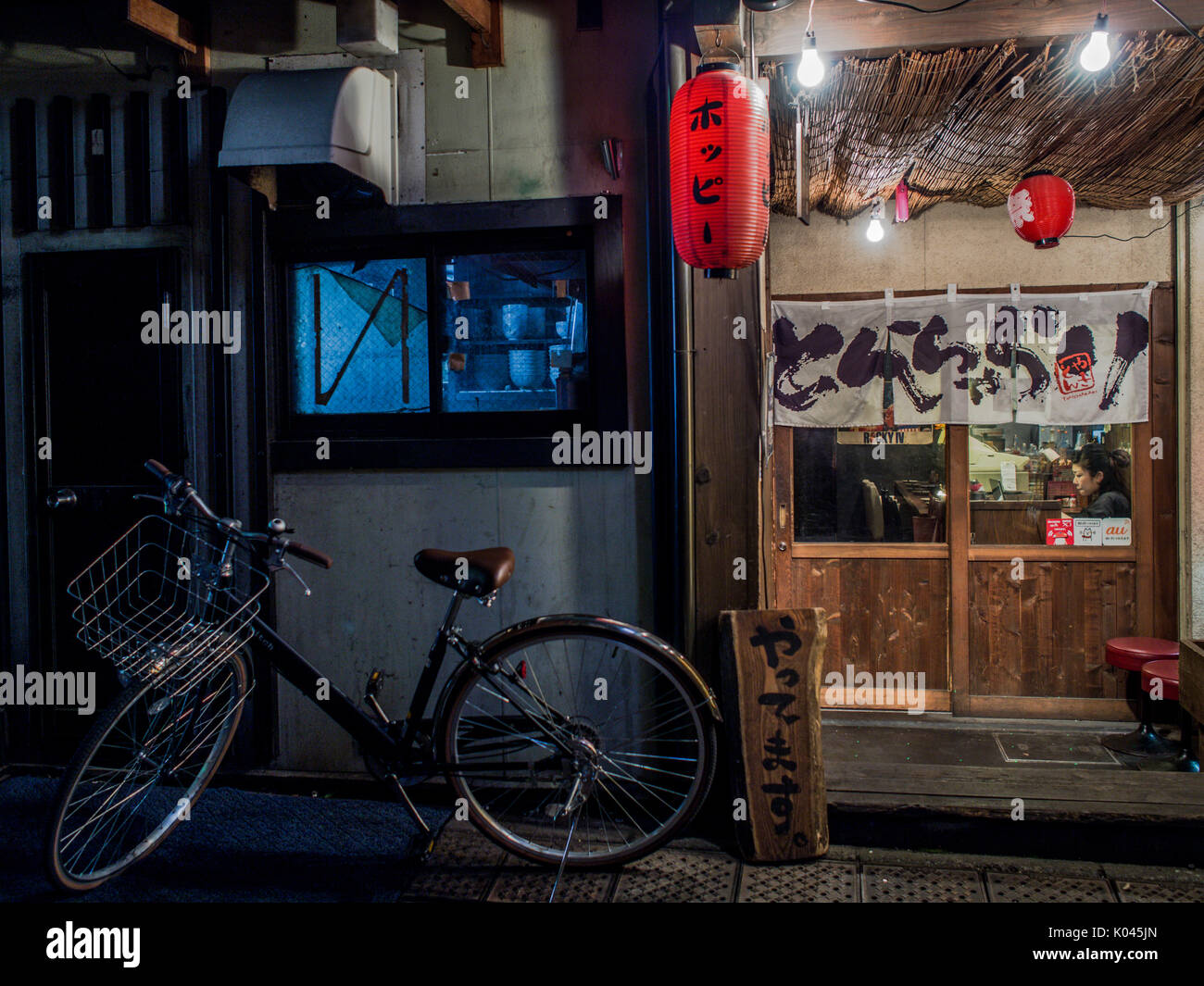 A bicycle parked outside a restaurant where a solitary woman sits alone. Night street, Ikebukuro, Tokyo, Japan Stock Photo